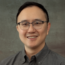 Chou Headshot - Romcorp Portrait of a man with short black hair and glasses, wearing a gray button-down shirt, smiling at the camera.