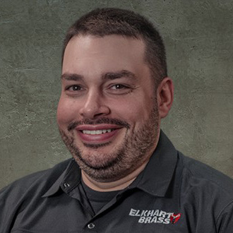 Dustin_Louden_square_ROM - Romcorp Portrait of a man with short dark hair and stubble, smiling, wearing a dark polo with the Elkhart Brass logo.